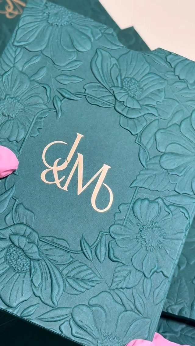 Teal invitation card with embossed flowers and gold foil-pressed initials J and M.