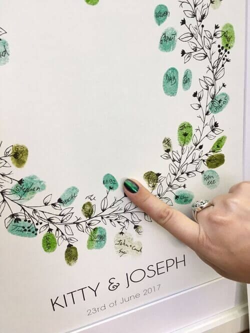 Hand pointing to a wedding guest book with green fingerprint leaves on a wreath.