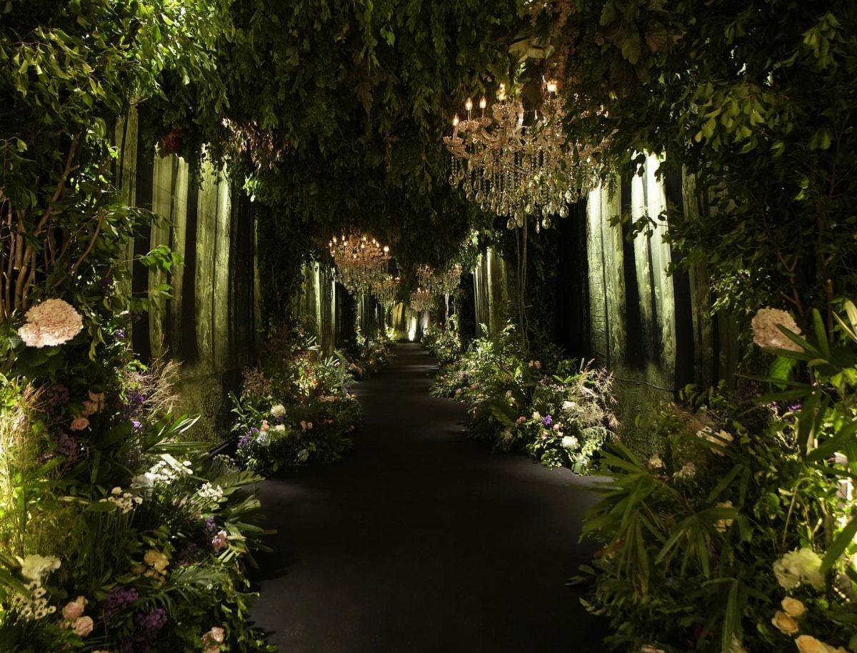 Lush garden walkway with crystal chandeliers, white flowers, and glowing vertical lights.