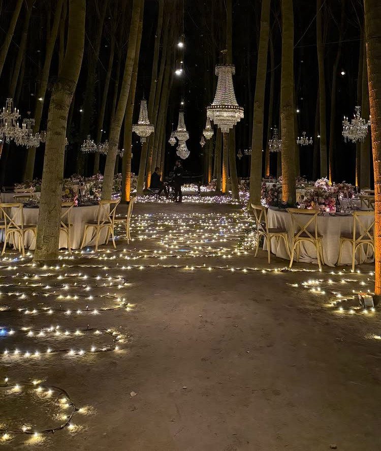 Crystal chandeliers hanging from tall forest trees above wedding tables and ground lights.