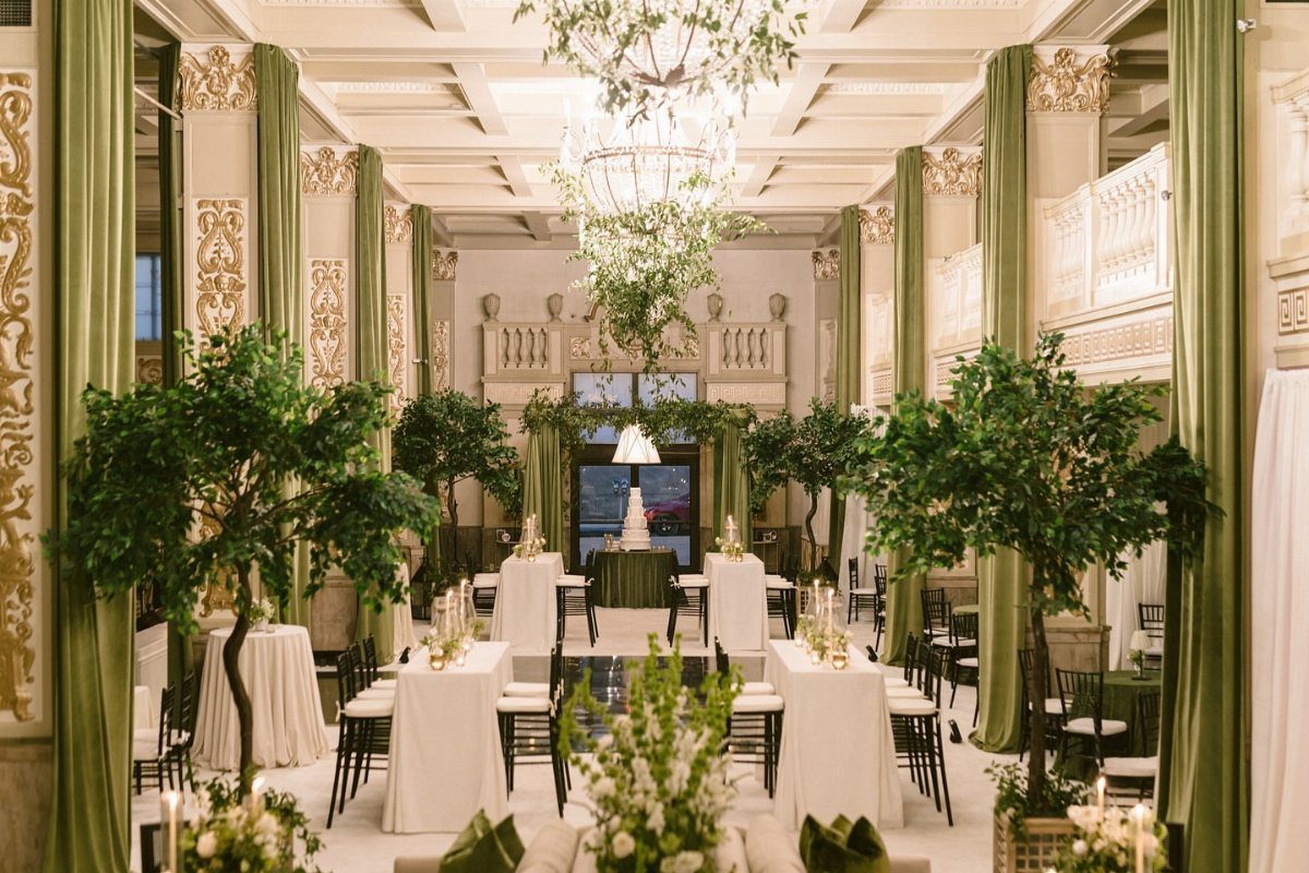 Ornate indoor hall decorated with potted trees, green curtains, and floral chandeliers.