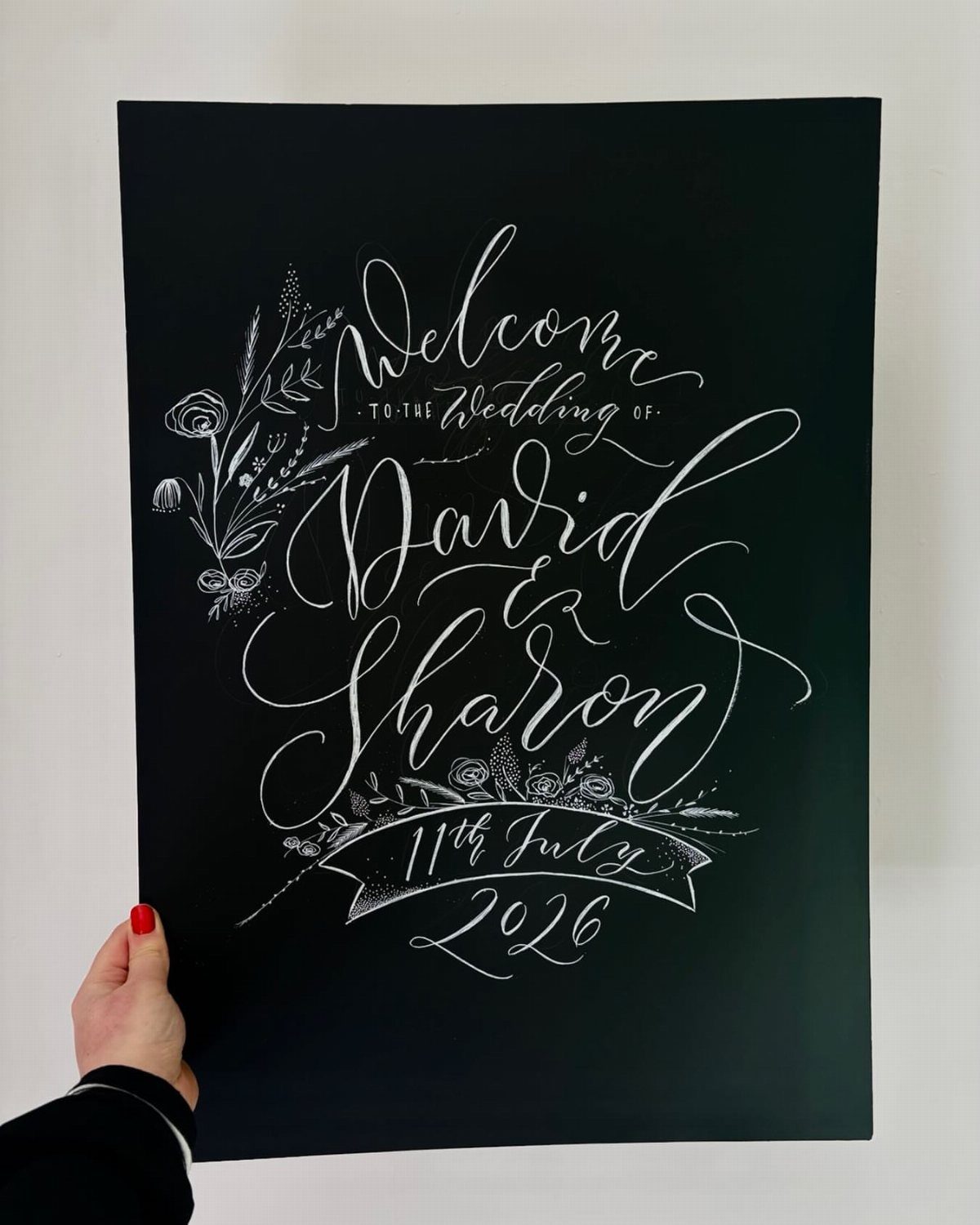 Black chalkboard wedding welcome sign with white calligraphy and floral accents held by hand.