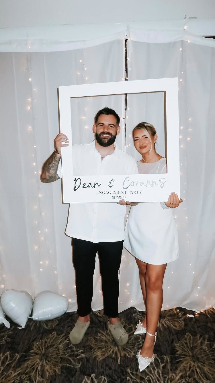 Couple holding a large white Polaroid-style frame around their faces in front of curtains.