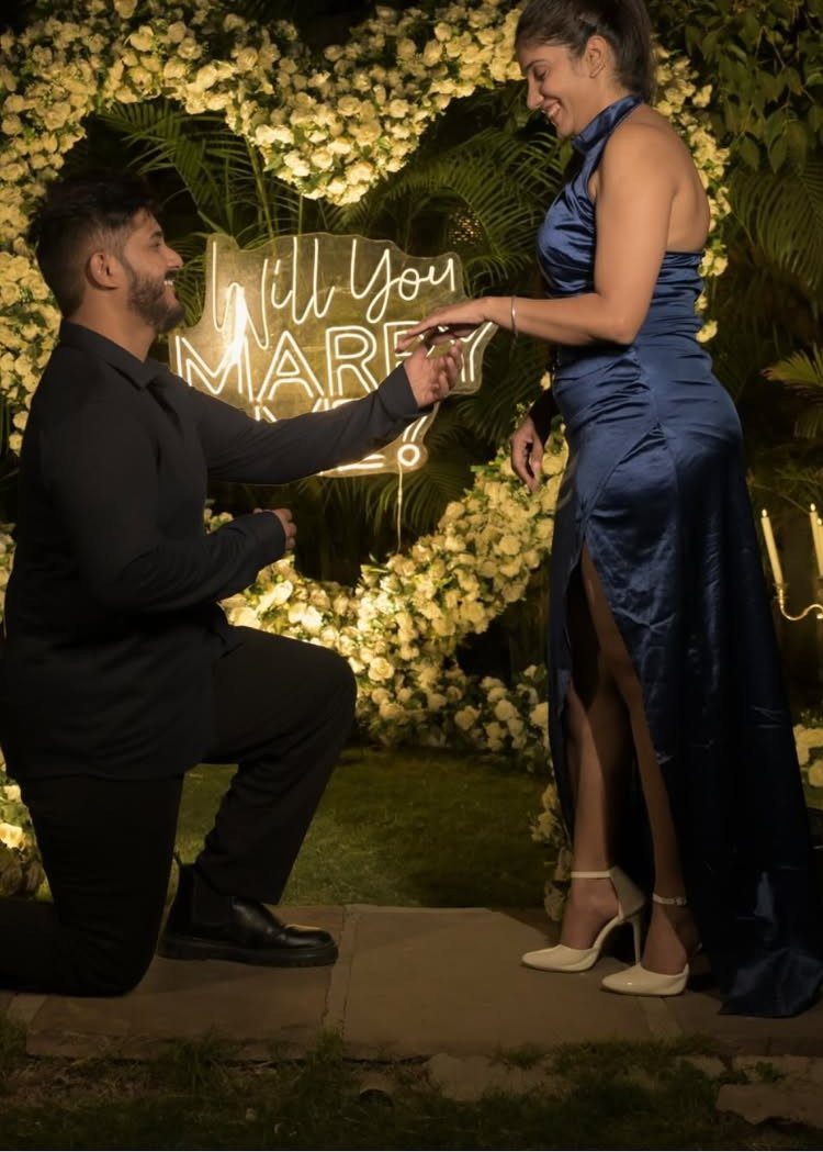 Man kneeling to propose to a woman in front of a floral heart wall.