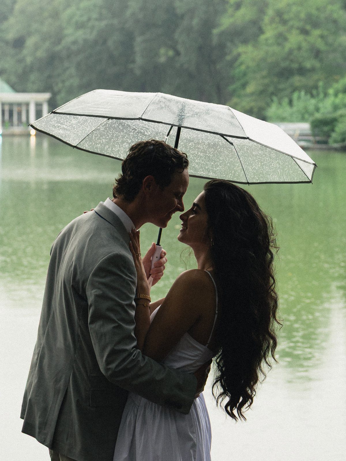 Couple embracing under a clear umbrella by a lake in the rain.