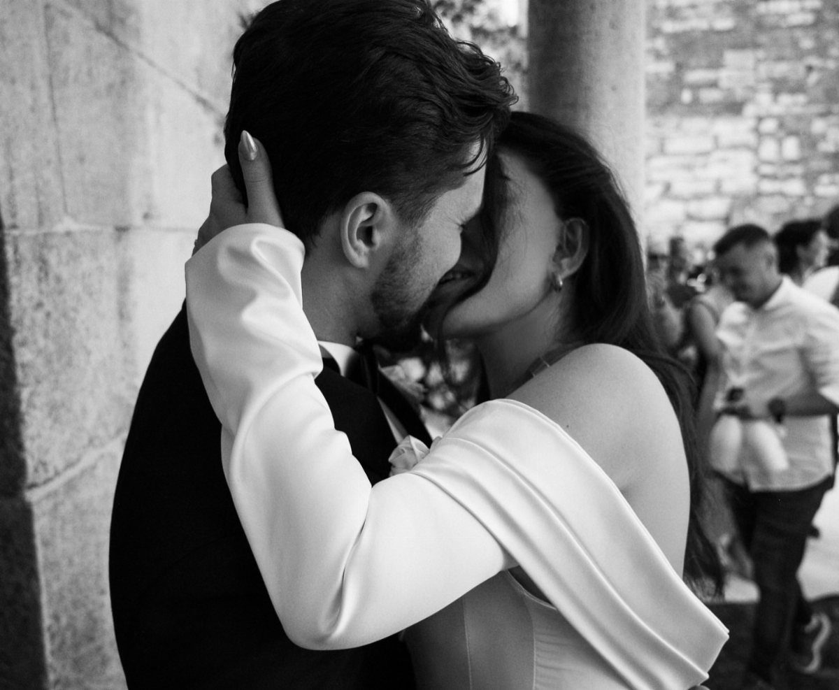 Couple in formal attire sharing a romantic kiss against a stone wall background.