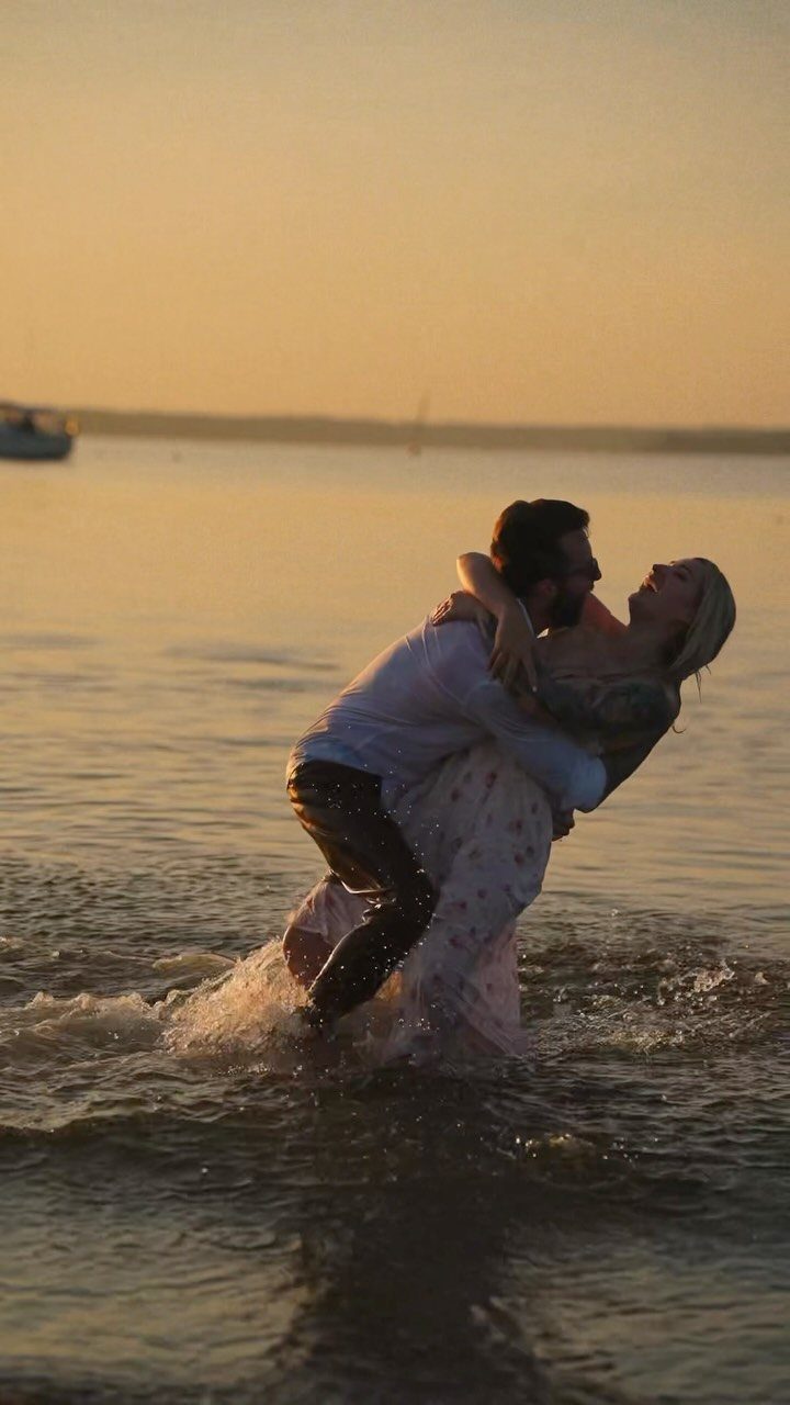 Couple embracing and dipping in shallow ocean water during a golden sunset.