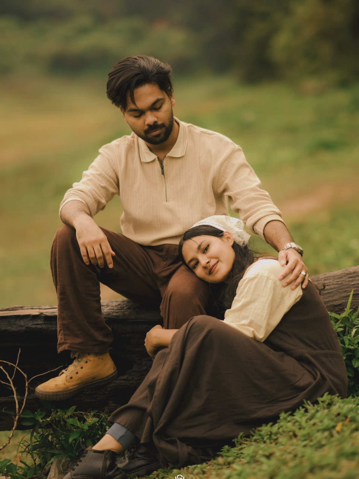 Couple in vintage-style beige and brown clothing posing on a fallen log outdoors.