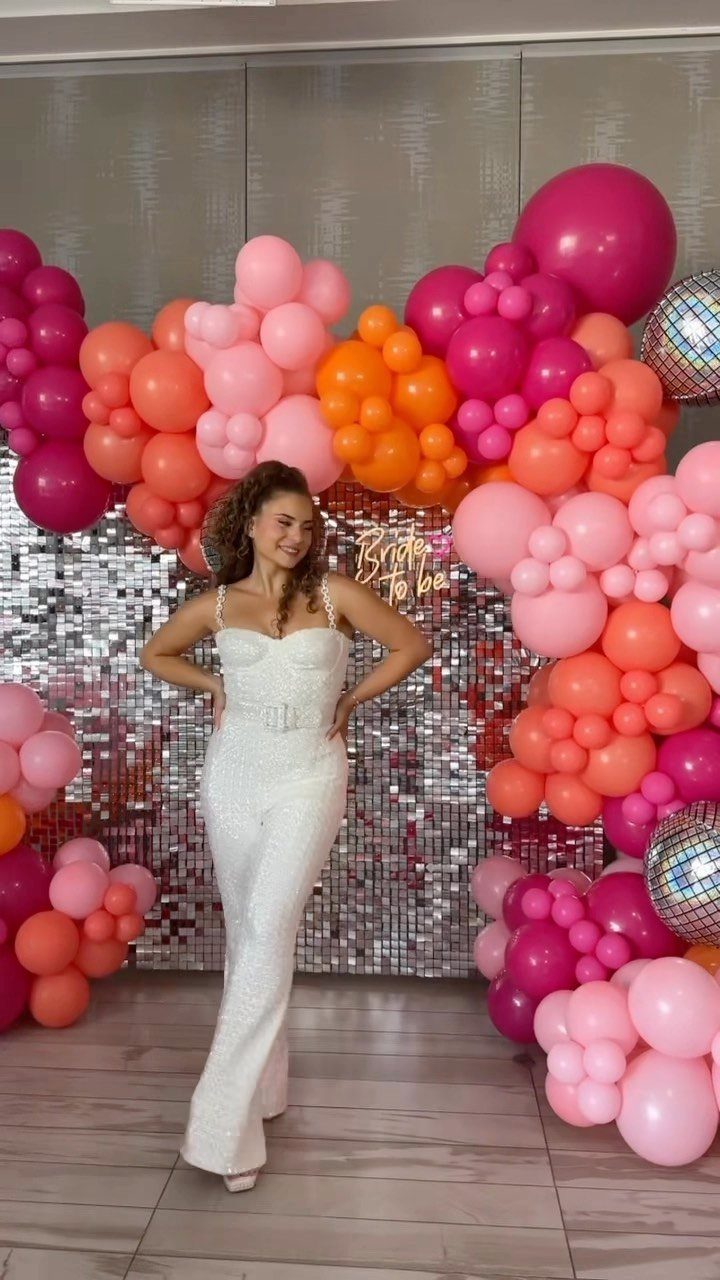 Woman in a white dressy jumpsuit posing before a pink balloon arch backdrop.