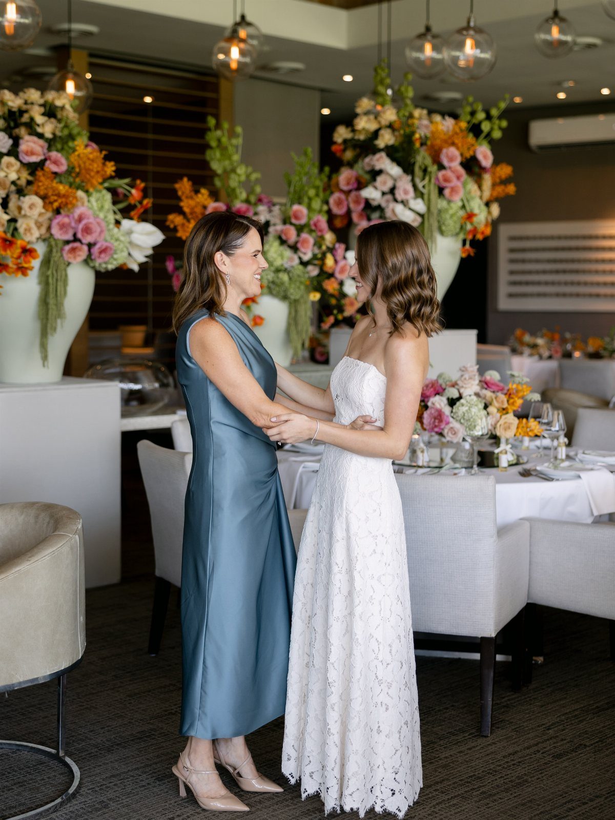 Two women in elegant dresses standing together in a room with floral centerpieces.