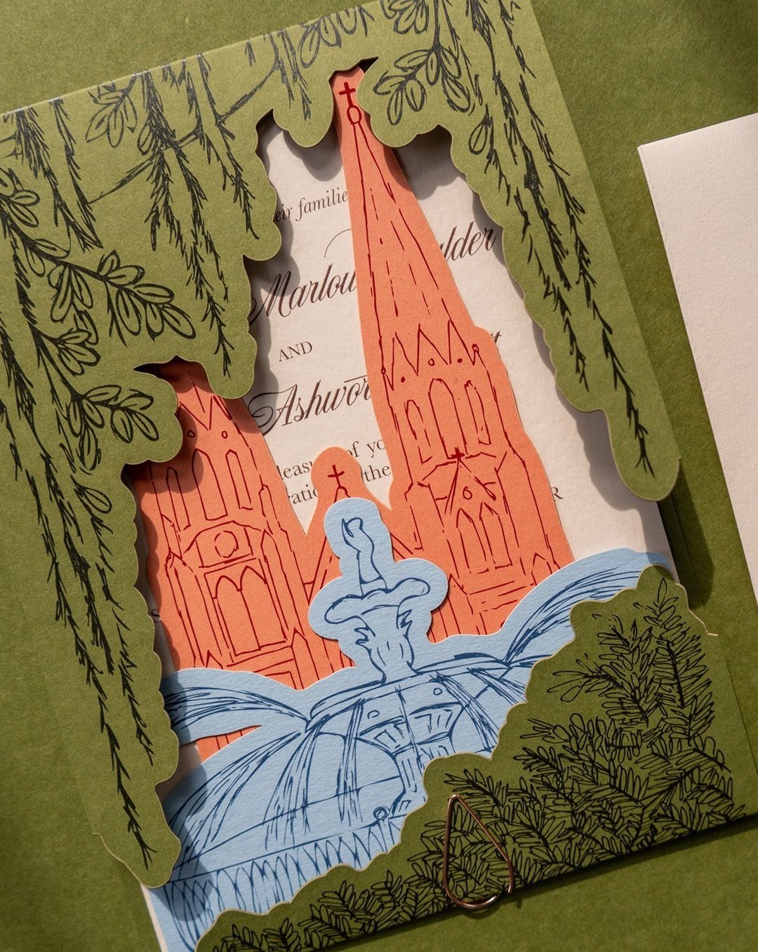 Layered wedding invitation with green laser-cut leaves, red cathedral, and blue fountain illustrations.