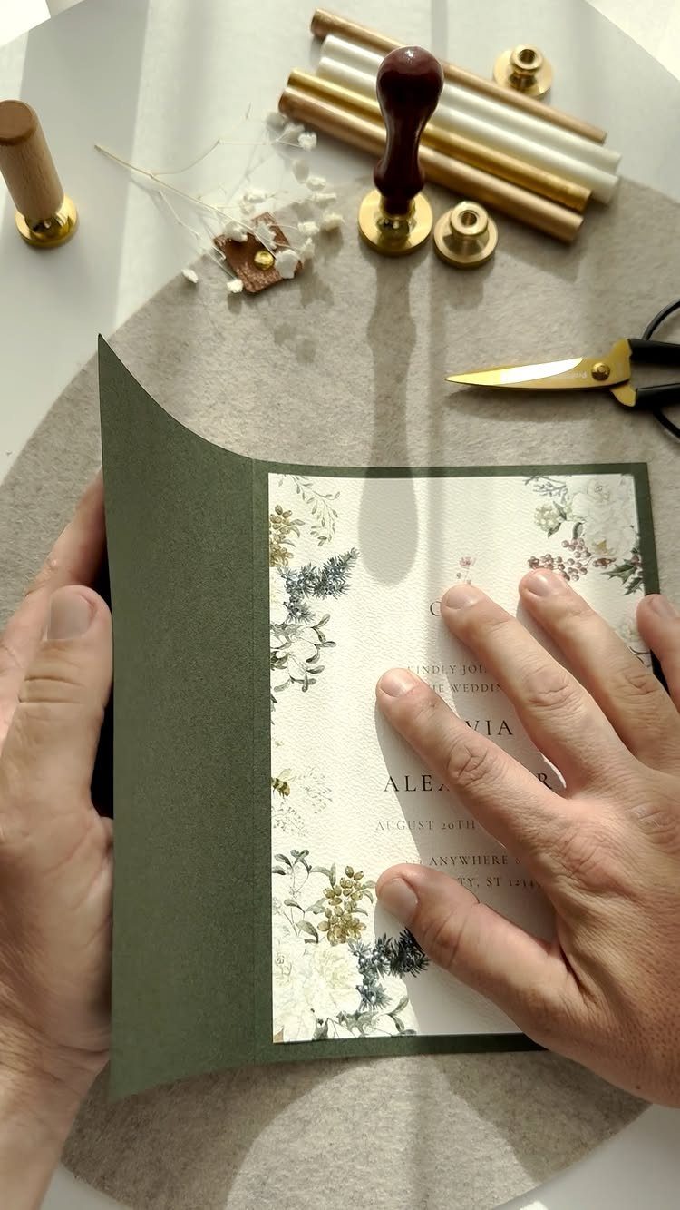 Olive green invitation folder with floral card, wax seals, and gold scissors.
