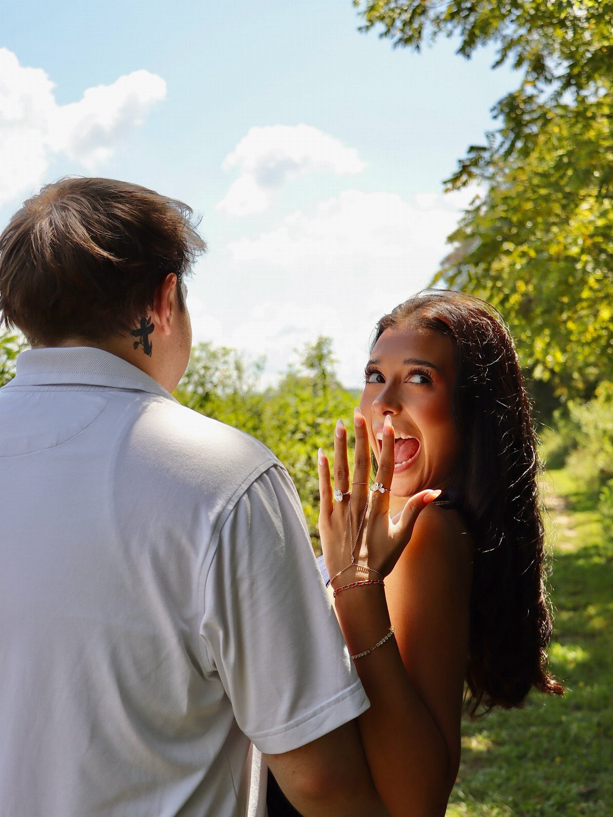 Excited woman showing her engagement ring to a man in a sunny outdoor setting.