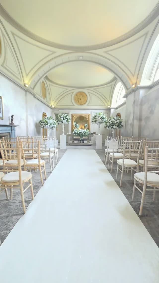 Wedding ceremony setup with white aisle runner, wooden chairs, and white floral arrangements.