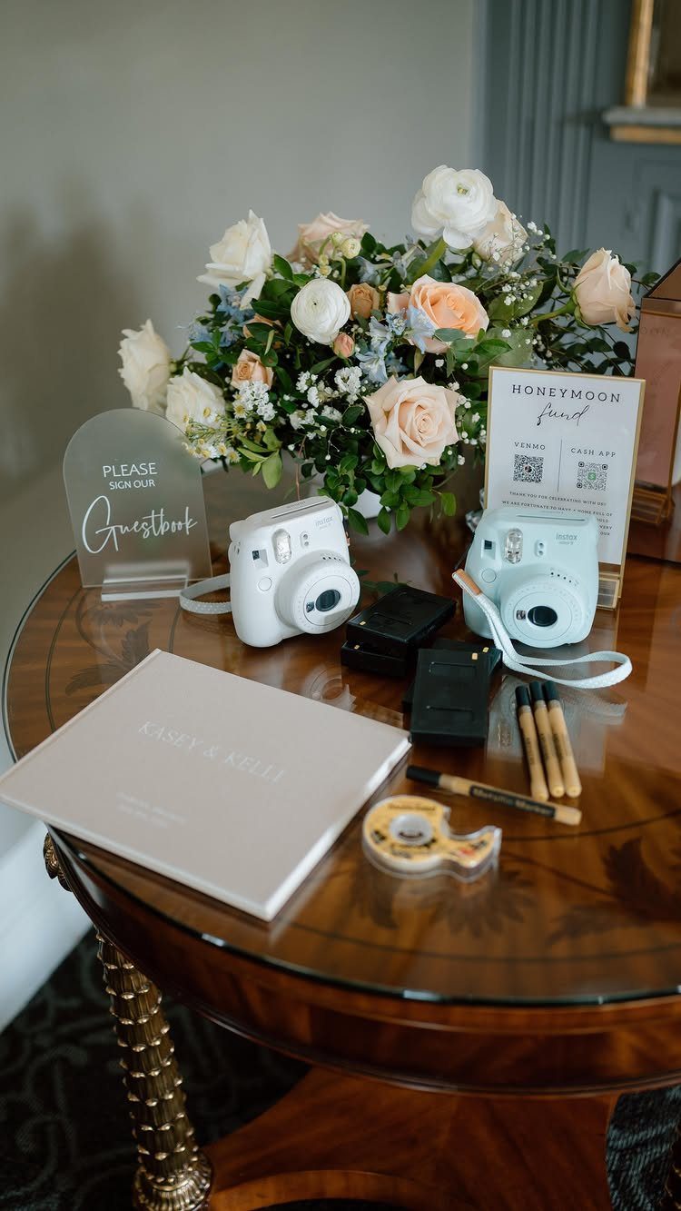 Instant cameras, a white guest book, and floral arrangement on a wooden table.