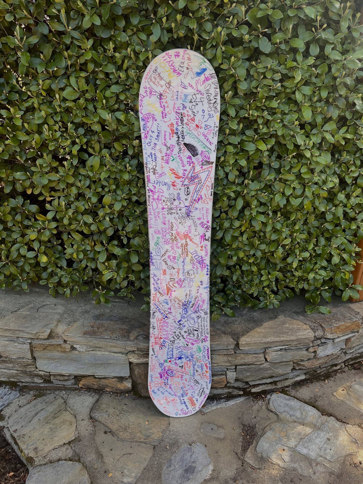 White snowboard covered in colorful handwritten signatures leaning against a stone wall.
