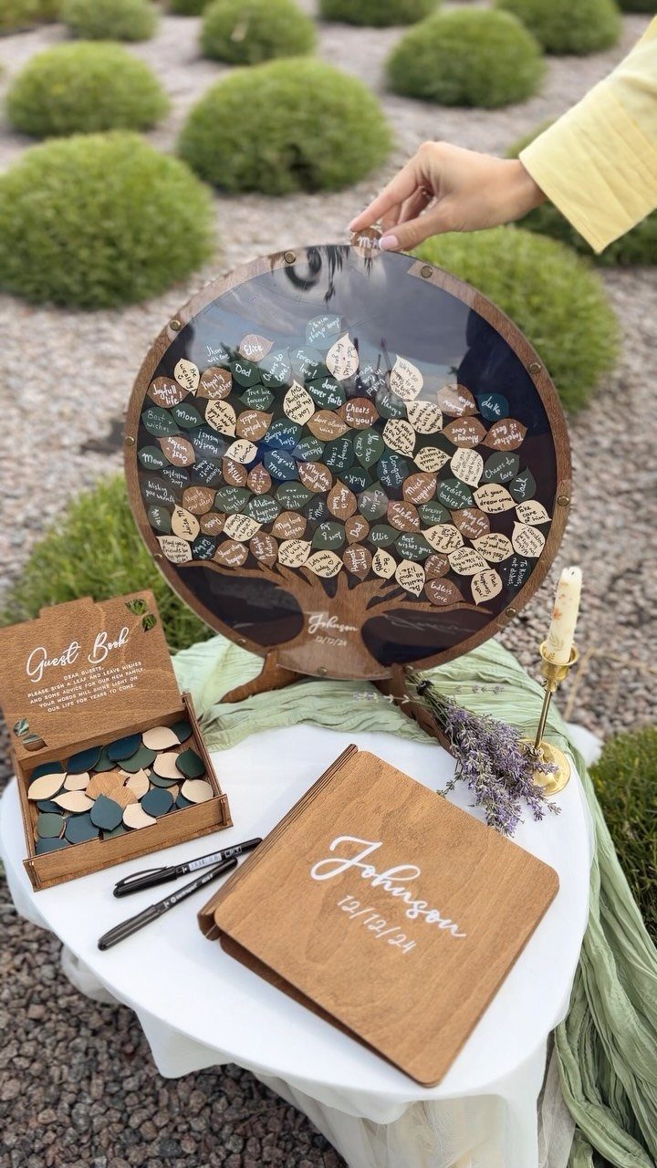 Circular wooden wishing tree with leaf tags and a personalized guest book outdoors.