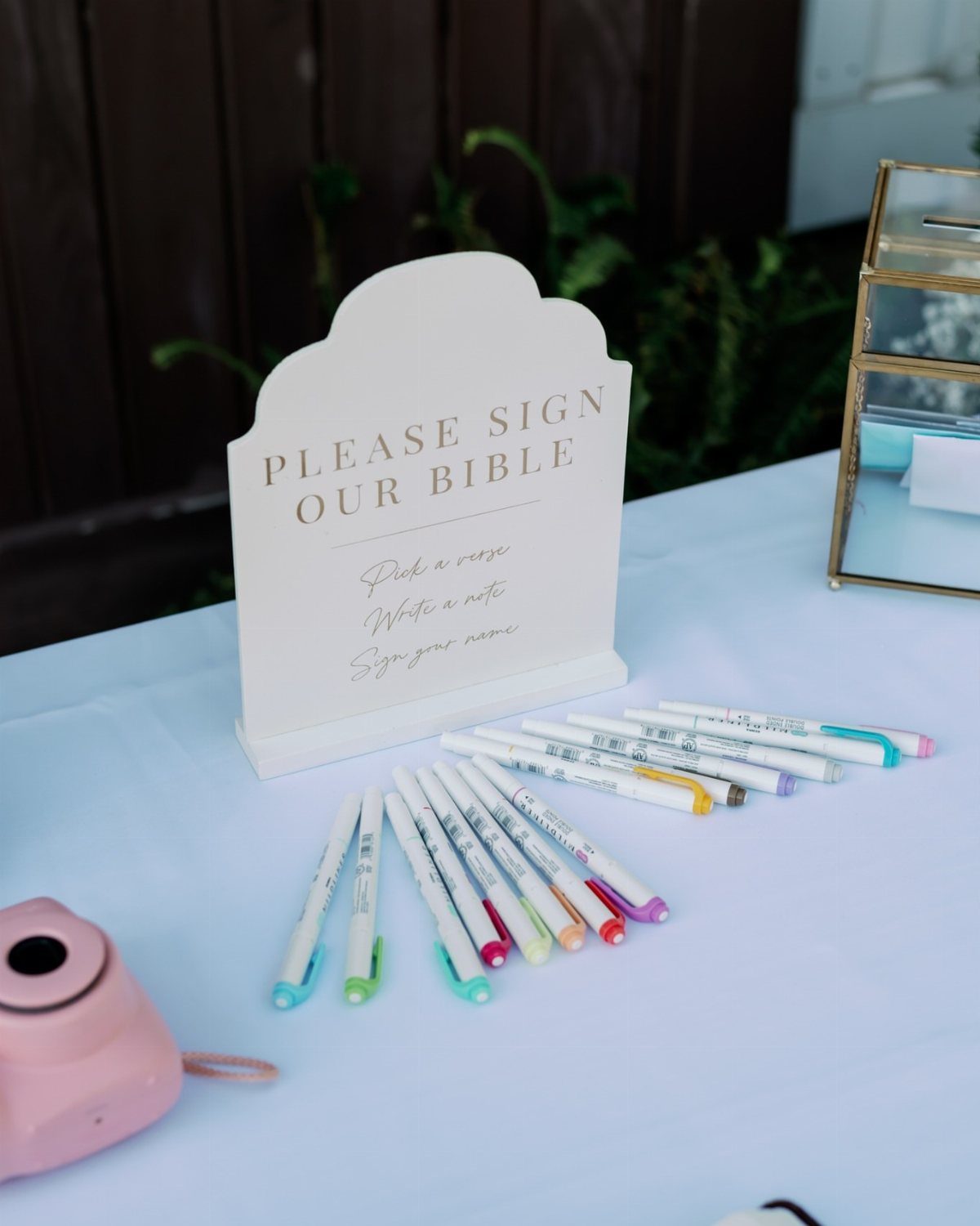 White arched sign requesting guests to sign a bible with colorful markers nearby.