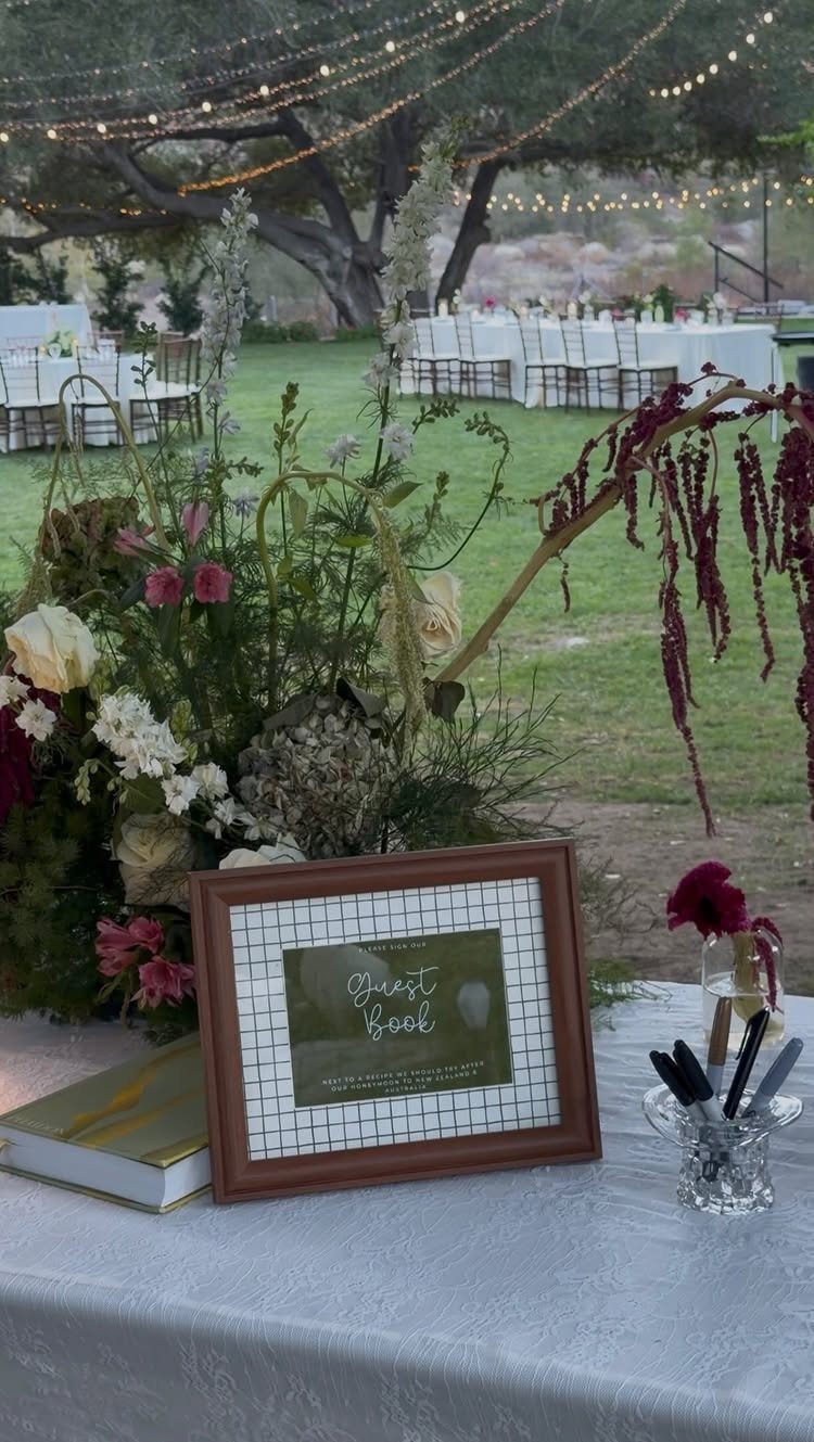 Framed guest book sign on a white table with flowers and pens outdoors.