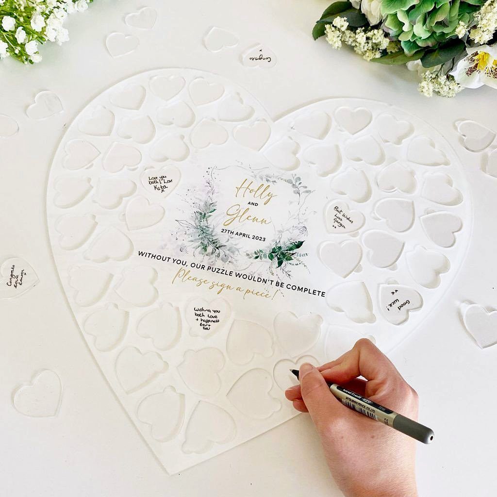 Hand signing a heart-shaped white puzzle guest book with a black marker.