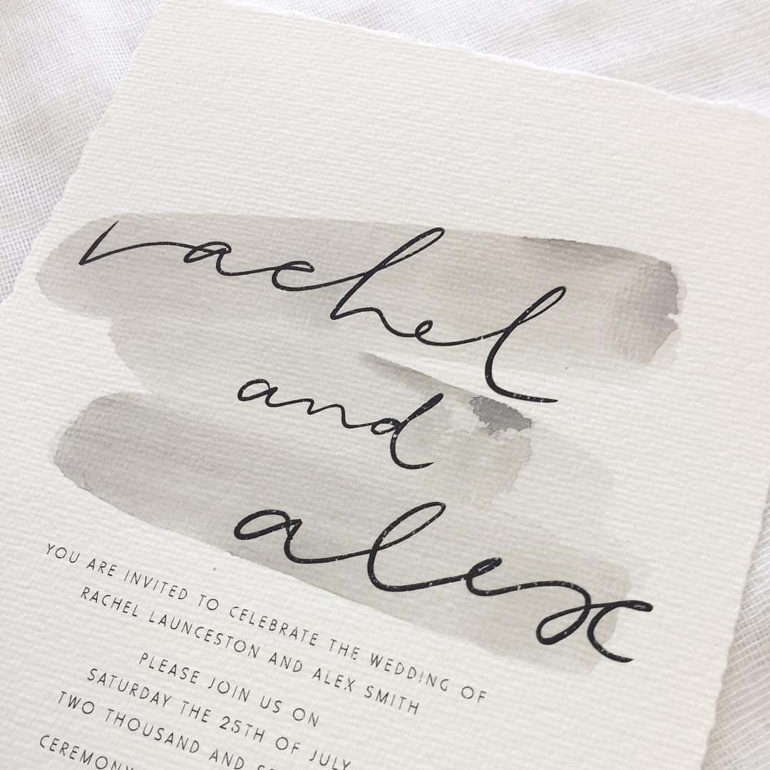 Wedding invitation with black calligraphy names over grey watercolor brushstrokes on white paper