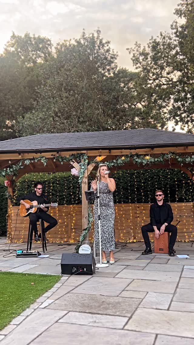Musical trio performing under a decorated wooden gazebo on a stone patio outdoors.