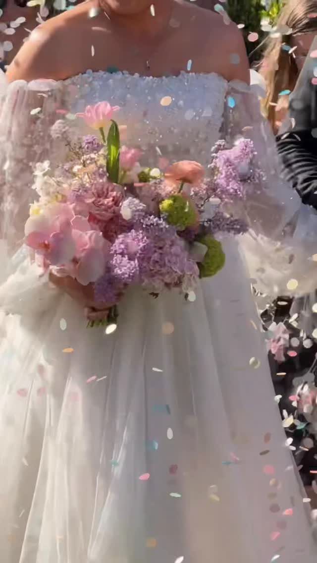 Bride in white dress holding a purple and pink bouquet with falling confetti.