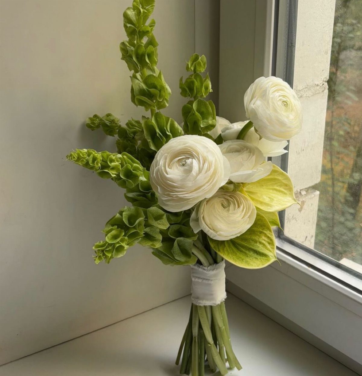 Bridal bouquet with white ranunculus and green foliage resting on a white windowsill.
