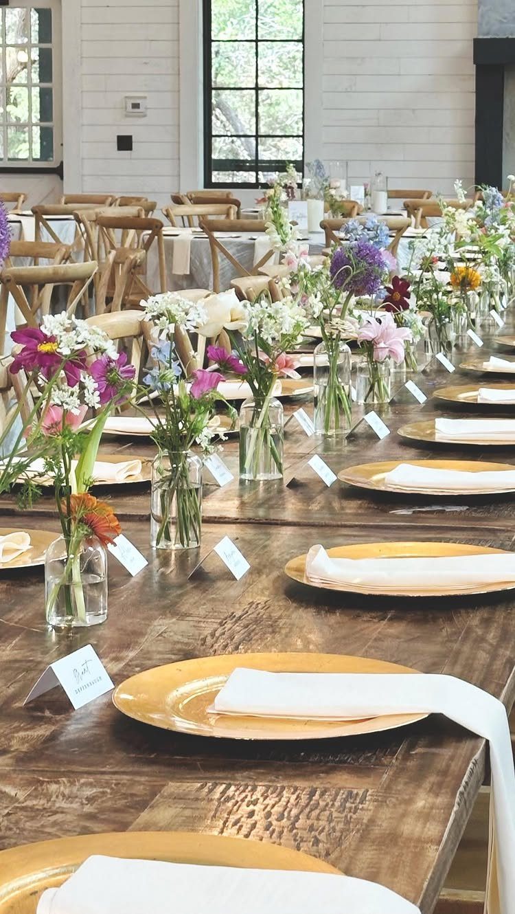 Wooden wedding table with gold plates, white napkins, and small wildflower arrangements.