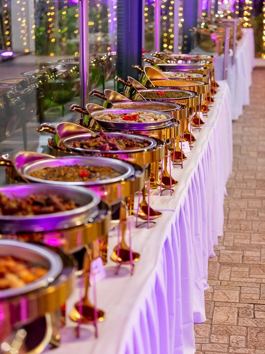 Buffet line of gold chafing dishes with food on a white tablecloth.