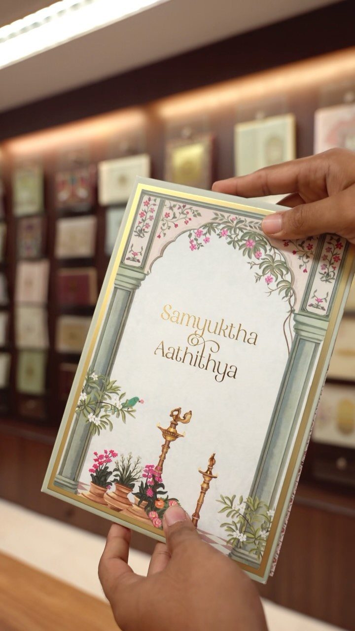 Indian wedding invitation card with floral designs and names held by two hands.