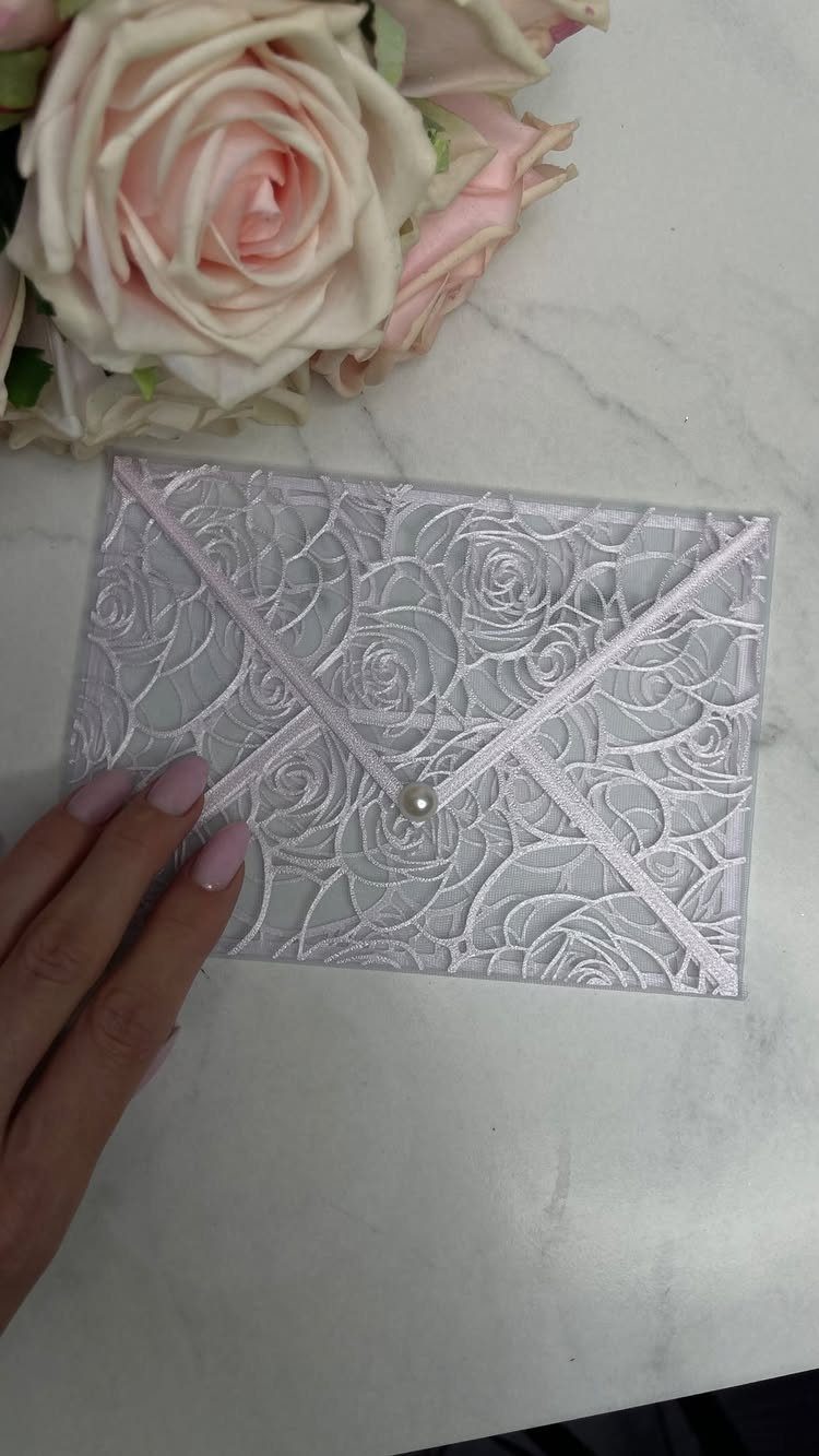 White laser-cut invitation envelope with rose patterns and a pearl closure beside pink roses.