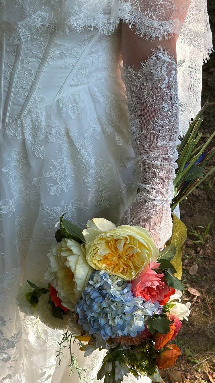 Bride in white lace dress and gloves holding a colorful floral bouquet.