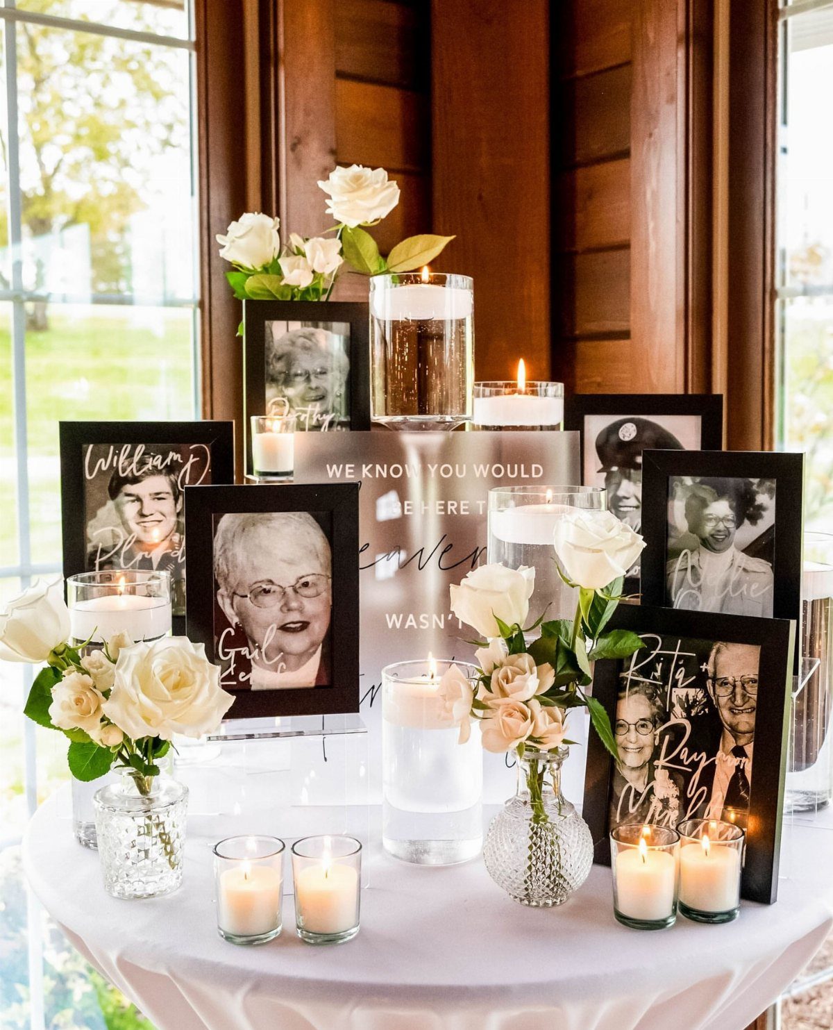Memorial table with framed portraits, white roses, and lit candles on white cloth.