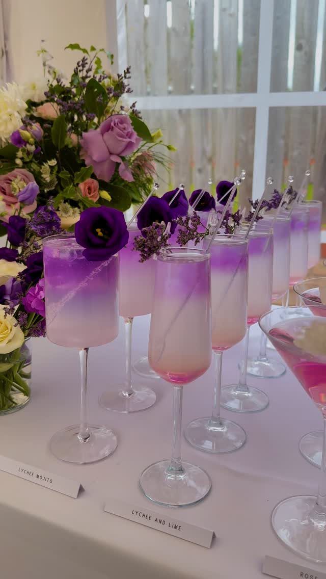 Layered purple and pink cocktails in champagne flutes next to a floral arrangement.