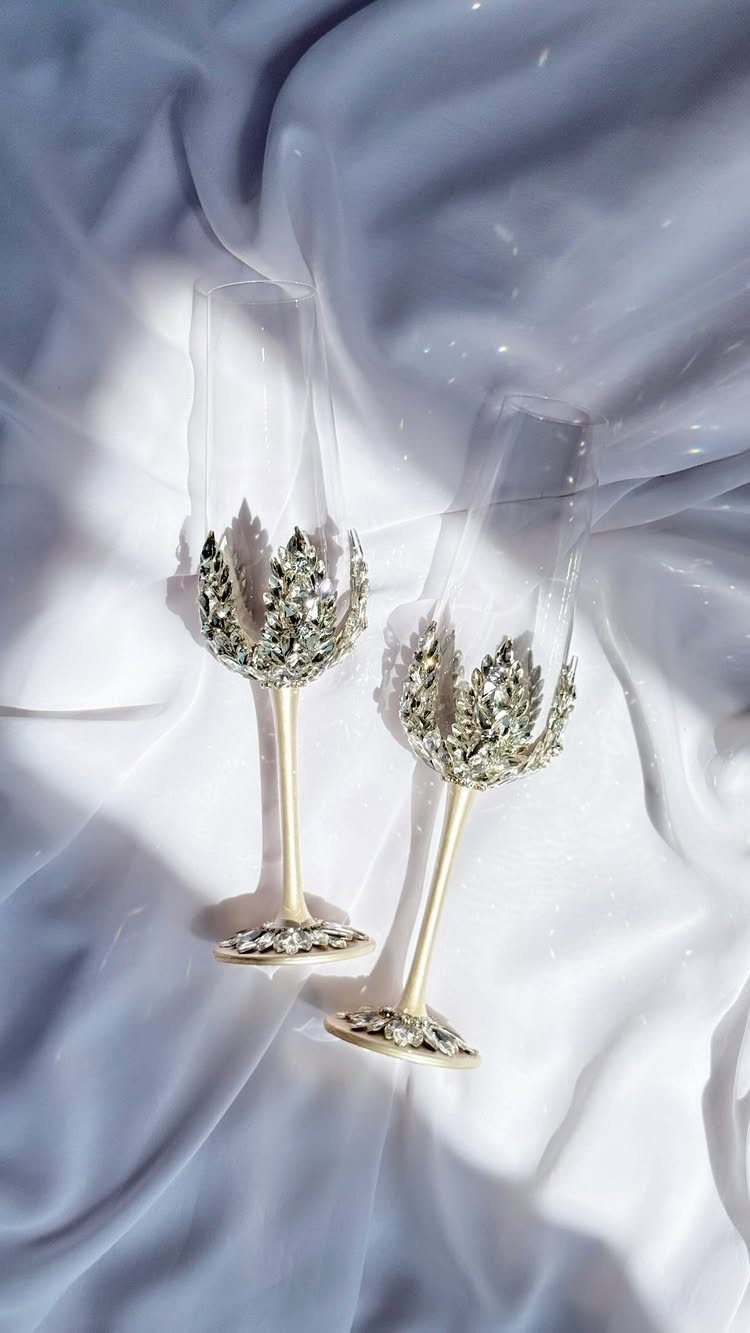 Two crystal-encrusted champagne flutes with gold stems on white satin fabric.