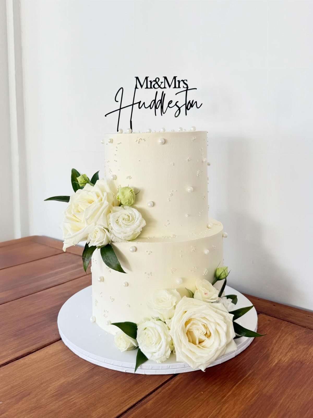 Two-tier white wedding cake with white roses and a black script cake topper.