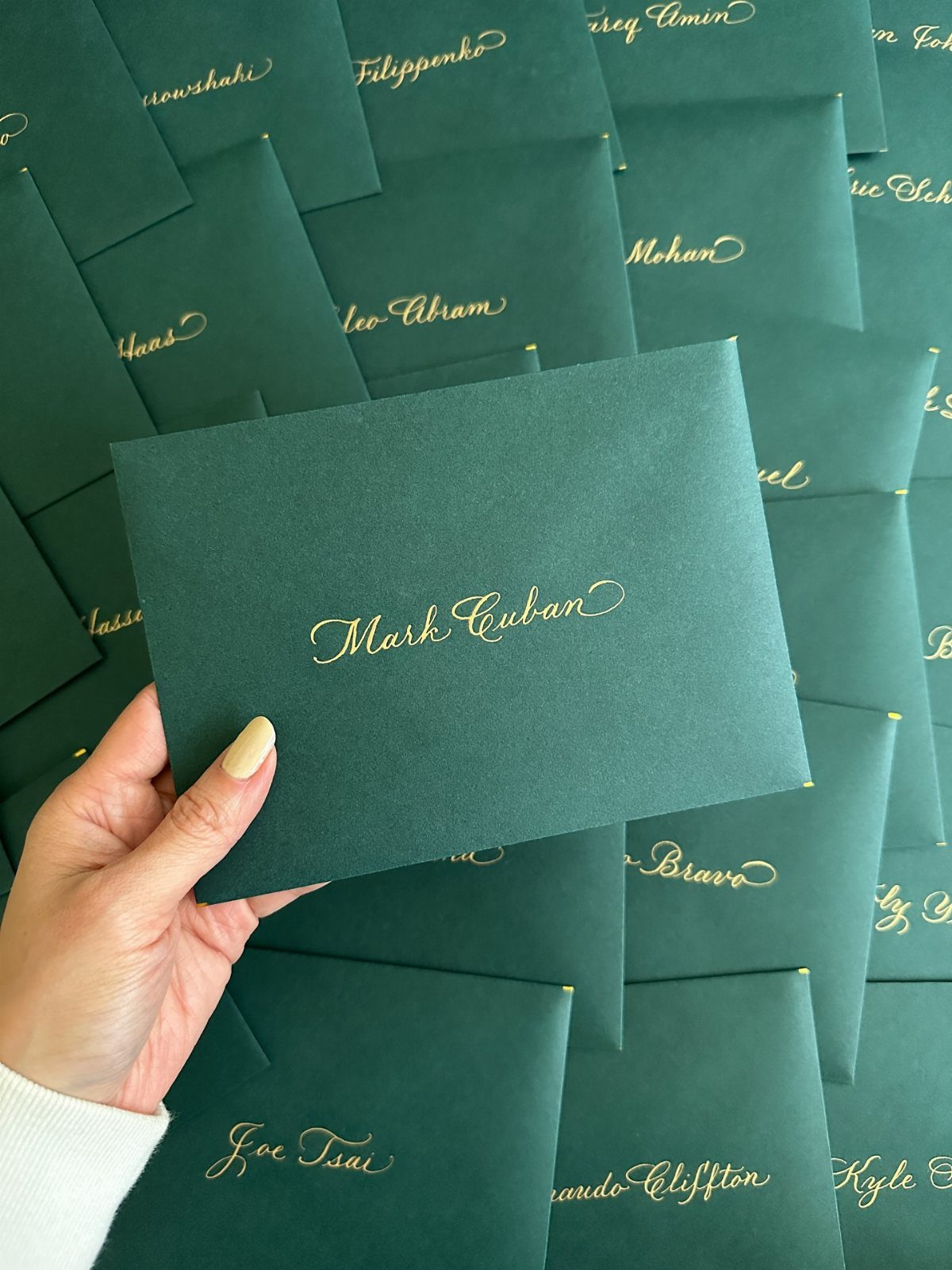 Dark green envelopes with gold hand-lettered calligraphy names held by a hand.
