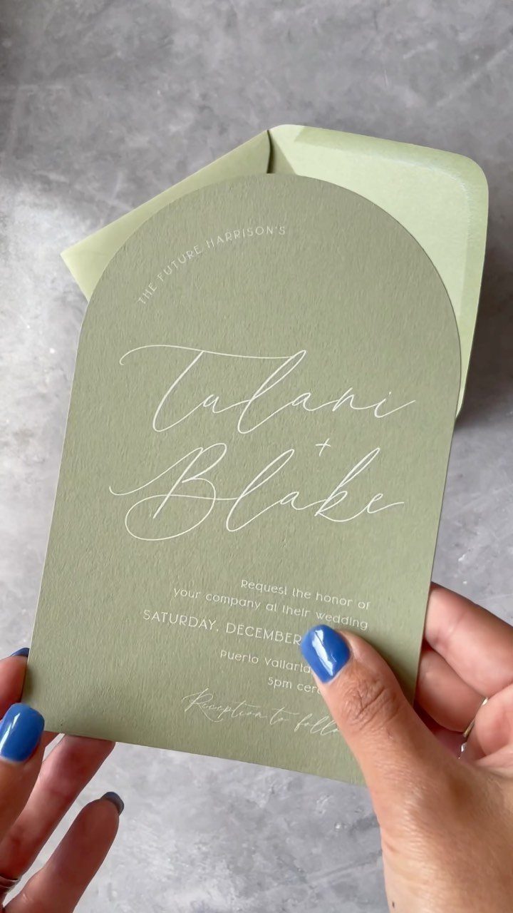 Sage green arch-shaped wedding invitation with white calligraphy held by blue-nailed hands.