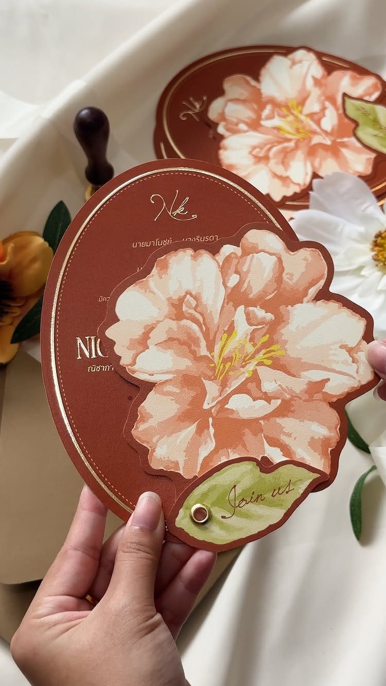 Circular reddish-brown wedding invitation with a large peach flower and gold calligraphy.