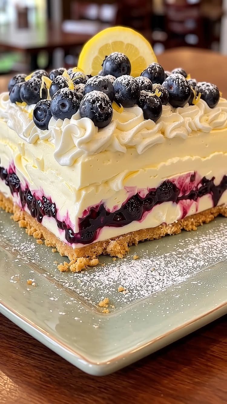 Layered cheesecake topped with fresh blueberries, whipped cream, and a lemon slice.