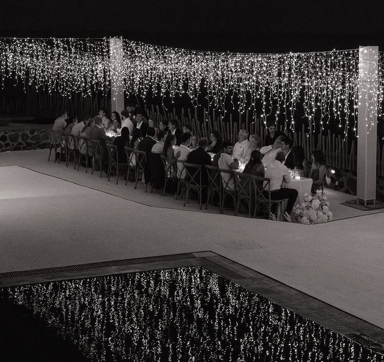 Guests dining at a long table under hanging fairy lights by a pool