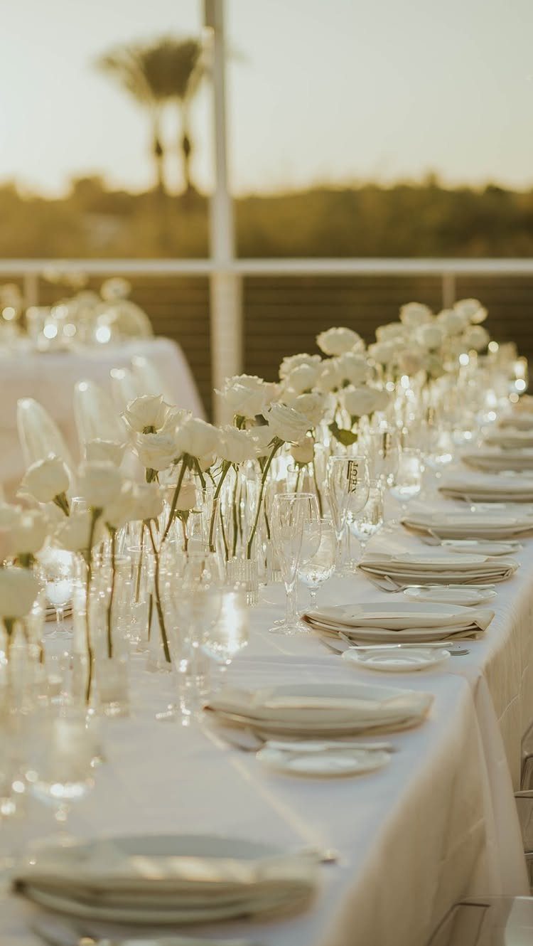 Long outdoor wedding table with white linens, plates, and white floral centerpieces.