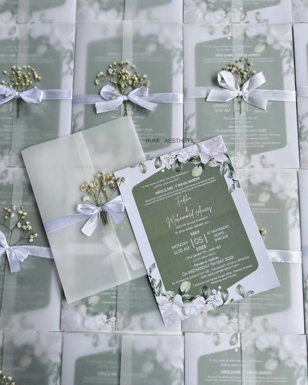 Sage green wedding invitations with white floral borders and vellum wraps tied with ribbons.