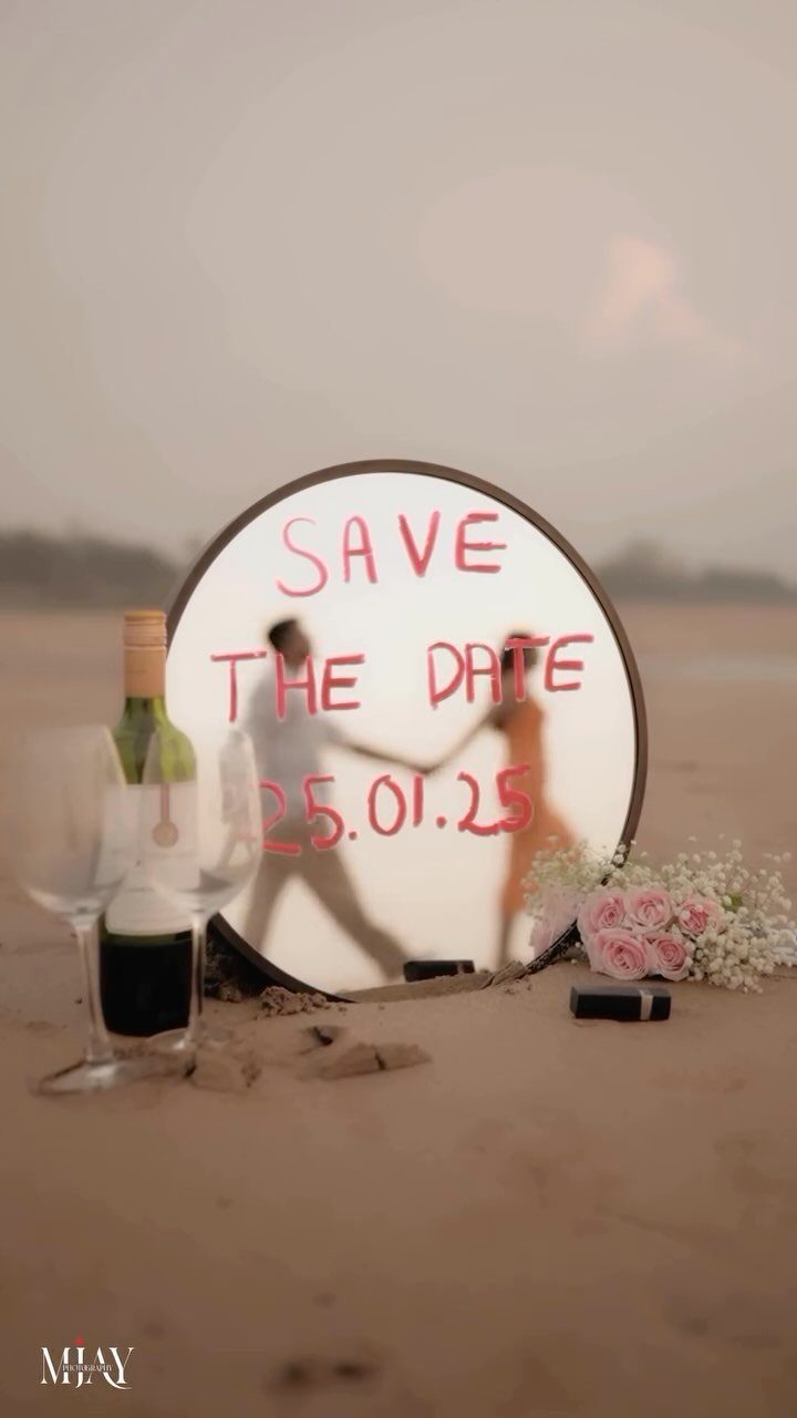 Circular mirror on a beach reflecting a couple with red save the date text