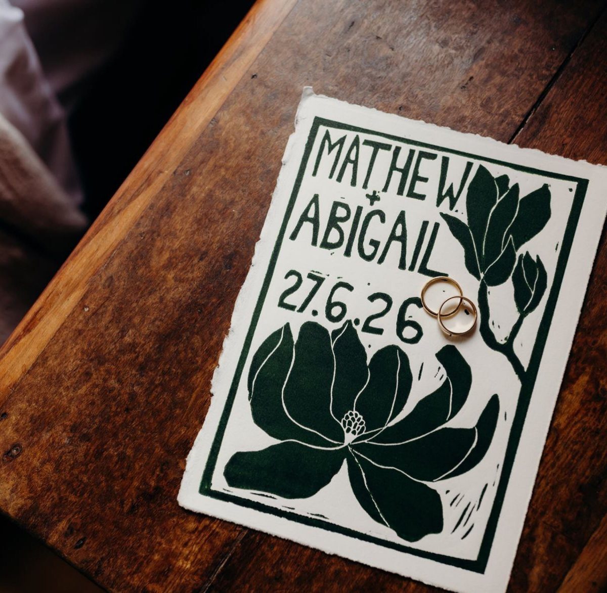 Save the date card with green floral art and gold rings on wood