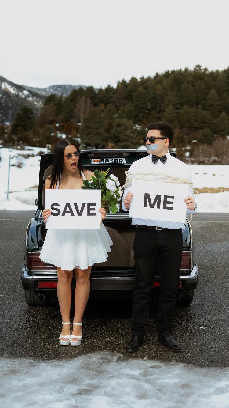 Couple posing by a car in snow, man tied up holding save me signs.