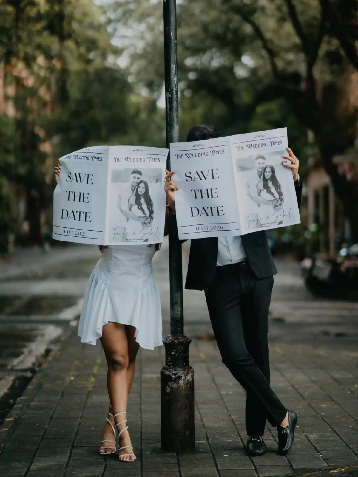 Couple holding newspaper-style save the date announcements covering their faces outdoors.