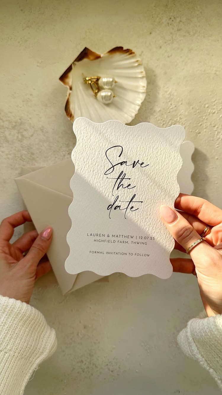Hands holding a white scalloped save the date card next to a seashell.