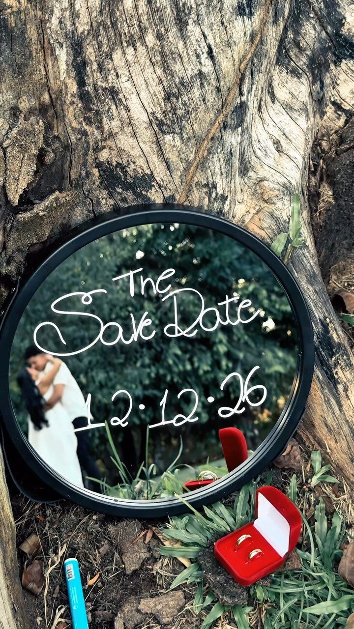 Circular mirror save the date with white script text resting against a tree trunk.
